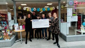 Staff of Aurora Managed Services and Shine A Light stand in front of their store with a Cheque